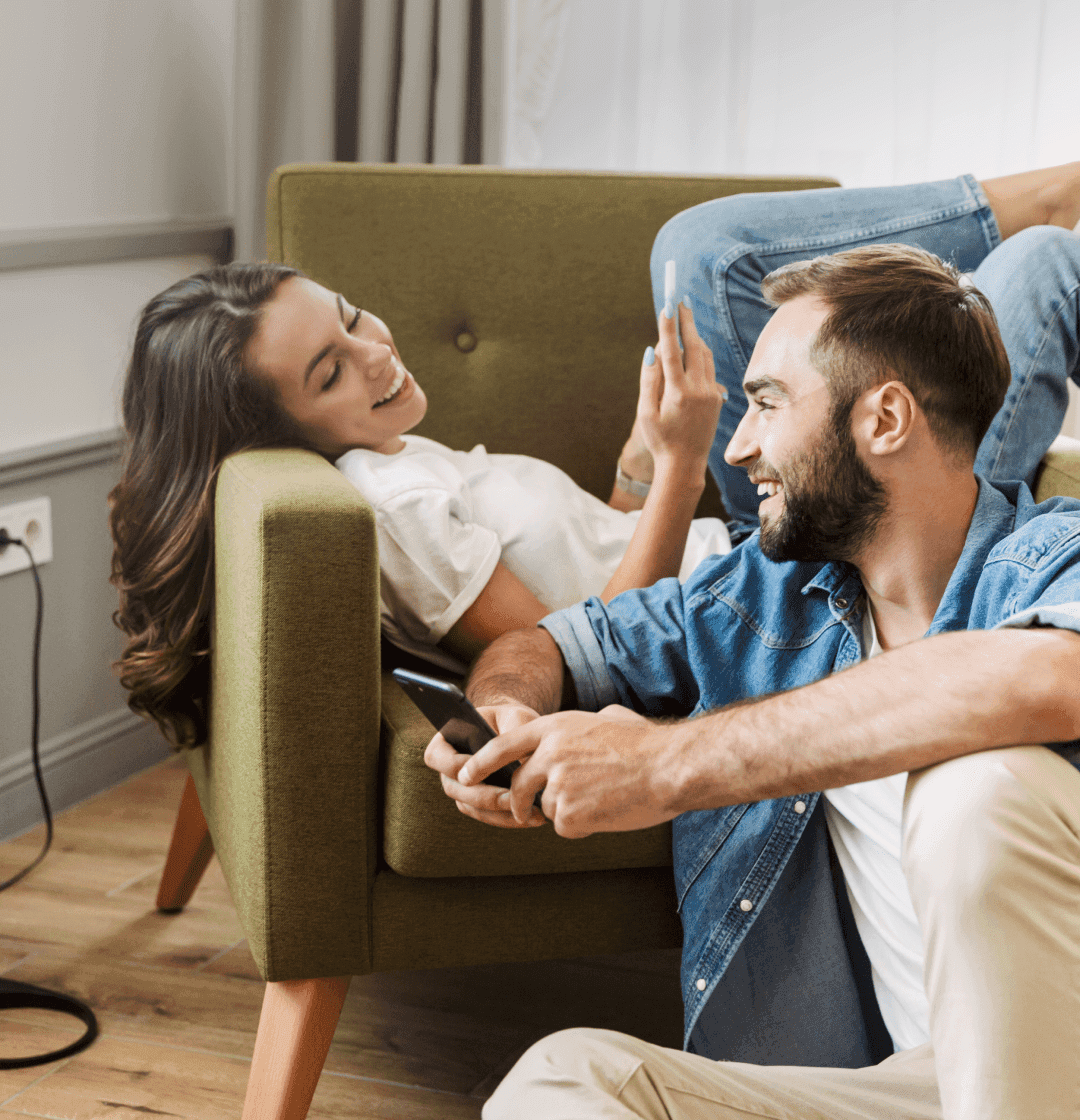 Una persona sonriendo acostada en un sillón con un celular en sus manos en conjunto de otra persona sonriendo sentada en el piso con un celular en sus manos